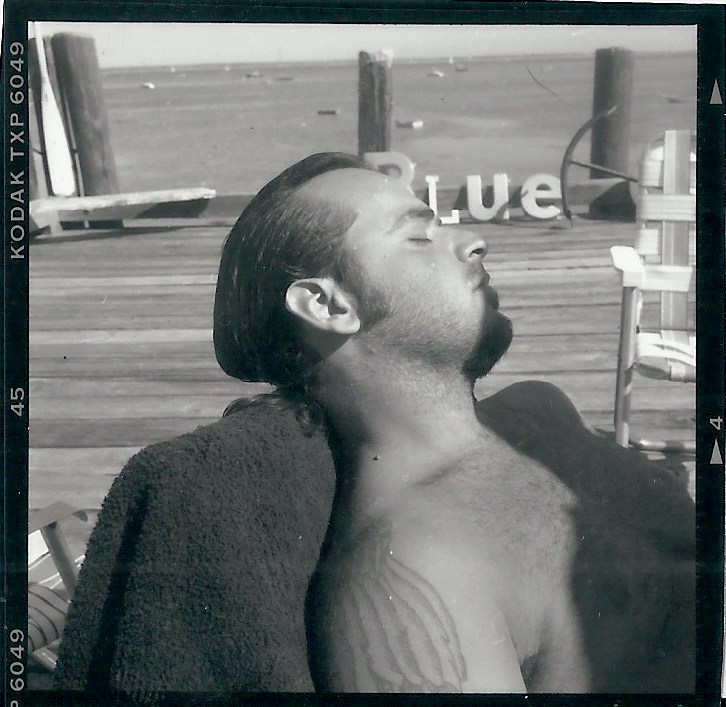 Portait of Guy Berube, Taken by David Carrino, at the Summer Residence of Jack Pierson, in Provincetown, Massachusetts. 'BLUE' word sculpture in background dock  by Pierson. Measures 2.25 x 2.25 inches. Private Collection. 